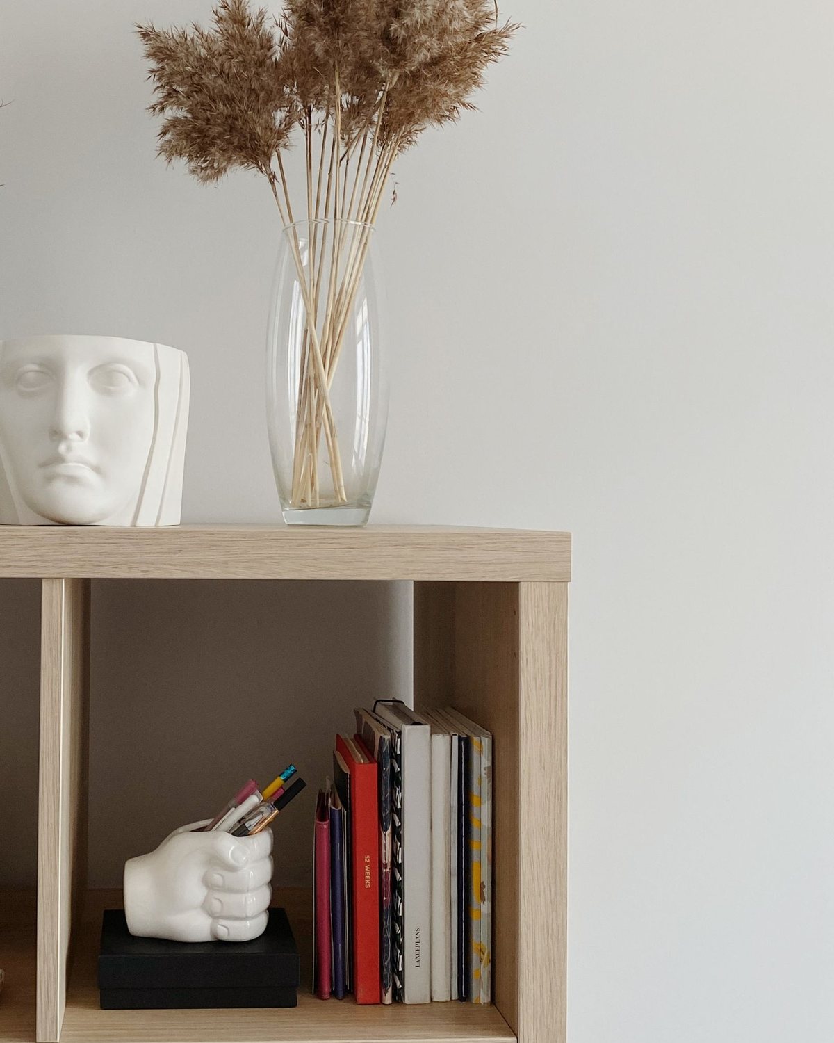 An oak-effect KALLAX shelf unit with a vase of pampas grass on top and books in a cube